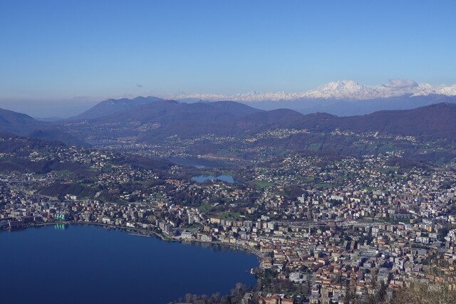 Über Lugano erheben sich die niedrigen Ketten vor dem Lago Maggiore, dahinter majestätisch und schneebedeckt die Walliser Alpen mit dem dominanten Monterosa-Massiv.