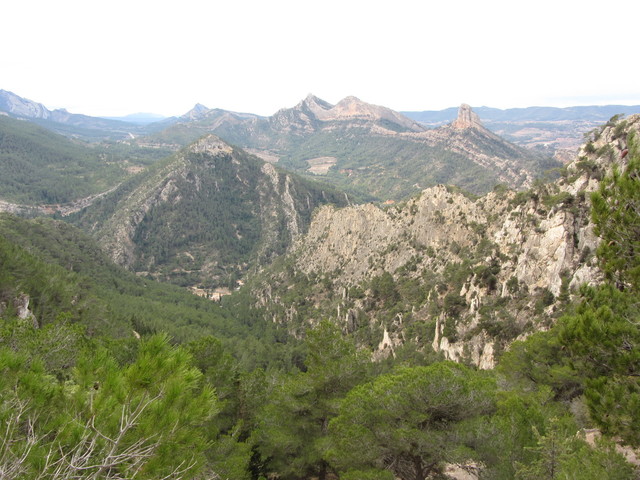 Südanfahrt: Blick vom Mirador am Ende des Steilstücks nach Süden. Zu erkennen ist das Santuari im Tal, die Via Verde dahinter und die Felsen Agulla de Bot, Falconera und Rocamala am Horizont.