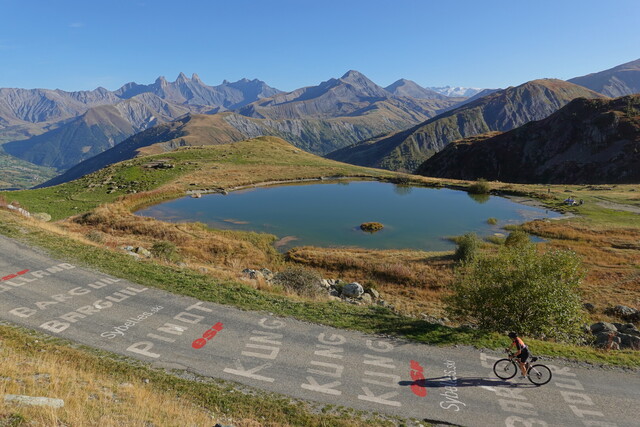 Schöner geht's kaum: schmale Straße ohne Verkehr, die Aiguilles d'Arves und der Meije-Gletscher im Hintergrund, mit Bergsee und Arvantal. Diesmal mit Radfahrerin.