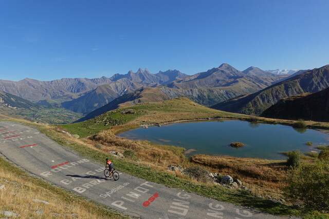 Schöner geht's kaum: schmale Straße ohne Verkehr, die Aiguilles d'Arves und der Meije-Gletscher im Hintergrund, mit Bergsee und Vallée de l'Arvan. Diesmal mit Radfahrerin.
