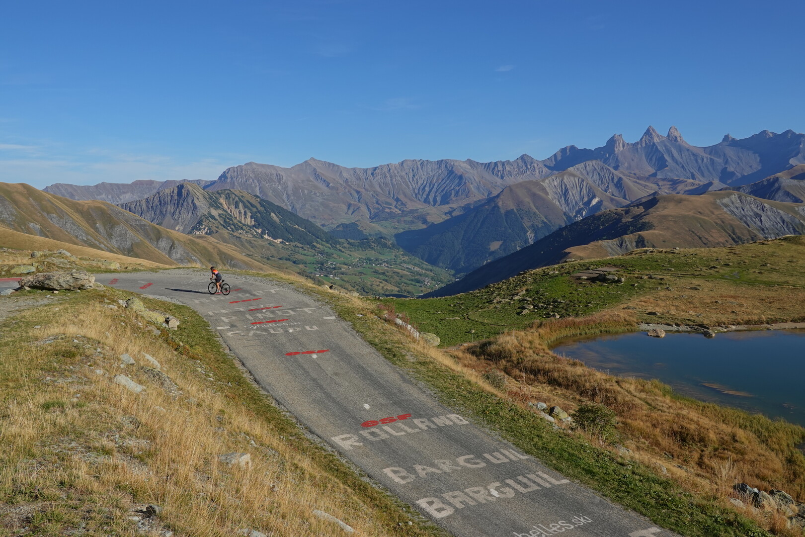 Schöner geht's kaum: schmale Straße ohne Verkehr, die Aiguilles d'Arves und das Vallée de l'Arvan im Hintergrund, mit Bergsee und Radfahrerin.